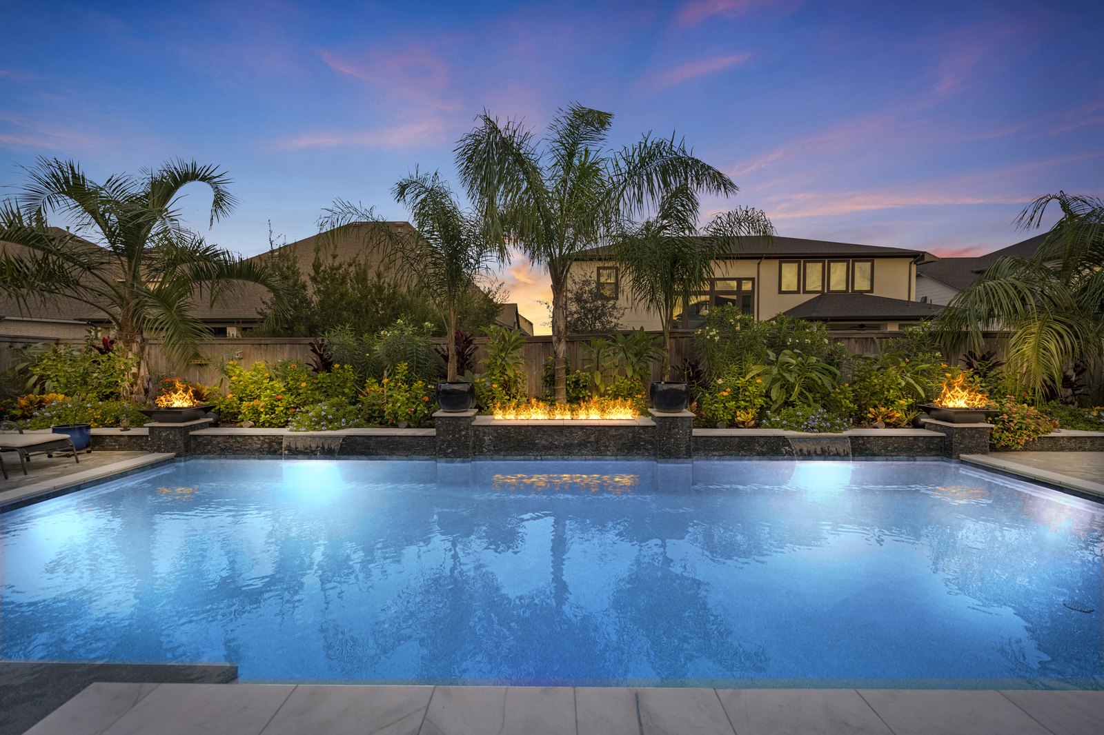 Virtual twilight of a palm-lined backyard pool with lit fire features